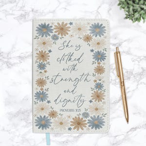May include: A floral-themed journal with the quote "She is clothed with strength and dignity" and the reference Proverbs 31:25. The cover features a daisy border in blue, tan, and cream colors. A gold pen rests beside the journal.