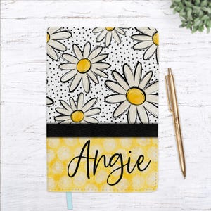 May include: A personalized notebook with a daisy pattern and the name "Angie" in cursive. The top half features white daisies on a black polka dot background, while the bottom half is yellow with a circular pattern. A gold pen rests beside it.