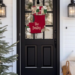 May include: A red Christmas stocking door hanger with the name "The Smiths" in white script. The top portion features a patchwork design with green, red, and beige squares. It hangs from a black door.