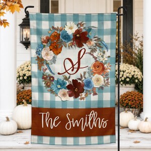 May include: A rectangular garden flag featuring a light blue and white buffalo check pattern. A central floral wreath in shades of orange, blue, and white surrounds a large script letter S. A solid brown horizontal band at the bottom reads The Smiths.