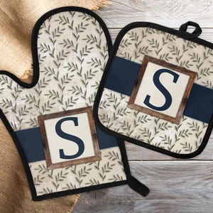 May include: An oven mitt and pot holder set with a cream-colored background and a leaf pattern. A navy blue band runs across each item, with a wooden-framed square displaying the letter "S". The set is trimmed with black.