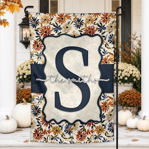 Könnte beinhalten: Eine rechteckige Gartenflagge mit einem Blumenmuster aus orangefarbenen, marineblauen und cremefarbenen Gänseblümchen. Eine mittige, cremefarbene Plakette mit Wellenrand zeigt ein großes marineblaues S und den kursiven Text the smiths.