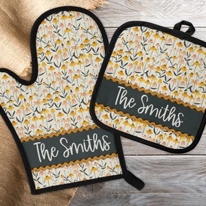 May include: A matching oven mitt and pot holder set. The set features a floral pattern with yellow and pink flowers on a cream background. A dark green banner with the text "The Smiths" is on each piece. Black trim.