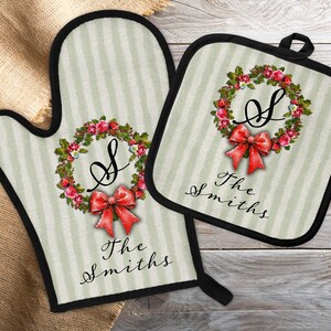May include: Oven mitt and pot holder set with a floral wreath design. The wreath features red roses and a red bow, with the letter "S" in the center. The text "The Smiths" is printed below the wreath. The set has a light green striped background and black trim.