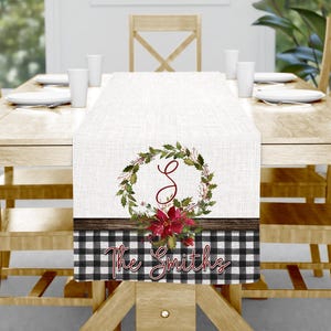 May include: A white table runner with a Christmas wreath design, a wooden border, and a black and white checkered pattern. The wreath features green leaves, red berries, and a red poinsettia. The text "The Smiths" is displayed.