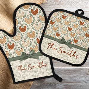 Könnte beinhalten: Ofenhandschuh- und Topflappen-Set mit Hühnermuster. Das Set hat einen cremefarbenen Hintergrund mit braunen und beigen Hühnern und grünen Blättern. Der untere Teil jedes Stücks hat ein grünes Band und den Text "The Smiths".