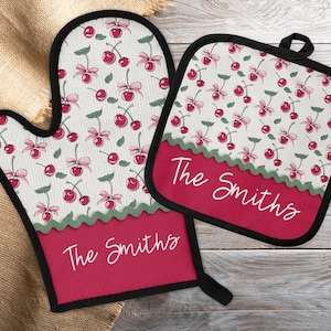 Puede incluir: Un juego de manopla y salvamanteles a juego. El conjunto presenta un fondo blanco con un estampado de cerezas rojas y lazos rosas. Una banda roja en la parte inferior de cada pieza tiene el texto "The Smiths" en escritura blanca. Ribete negro.