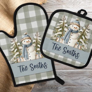 Puede incluir: Un juego de manopla y agarradera a juego. El conjunto presenta un tema invernal con un muñeco de nieve con gorro y bufanda azules, sobre un fondo de árboles nevados y regalos. El texto "The Smiths" se muestra en una pancarta azul.