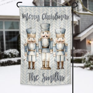 Puede incluir: Una bandera de jardín con un fondo de patrón de chevrón azul claro y blanco. La bandera presenta tres figuras de cascanueces de estilo acuarela con uniformes azules y blancos. El texto "Merry Christmas" y "The Smiths" están escritos en la bandera.