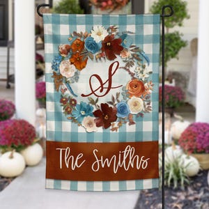 May include: A garden flag with a blue and white checkered background. It features a floral wreath with the letter "S" in the center. The bottom has a brown banner with the words "The Smiths" in white script.
