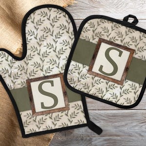 May include: Oven mitt and pot holder set with a beige background and a green leaf pattern. Each piece has a wooden-framed square with the letter "S" in the center. The set is trimmed with black and has a green band across the middle.