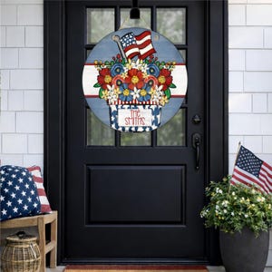 Pode incluir: Uma placa de porta redonda de madeira com um arranjo floral patriótico, apresentando flores vermelhas, brancas e azuis e duas bandeiras americanas. A placa exibe o texto "THE SMITHS", perfeita para receber convidados.