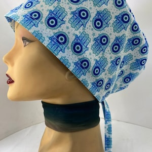 May include: A white surgical scrub hat with a blue and white pattern of hamsa hands and evil eyes. The hat has a tie at the back.