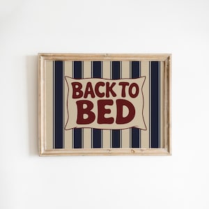 May include: A framed print featuring the phrase "BACK TO BED" in a retro style. The text is displayed on a beige pillow-shaped design, set against a background of navy blue and beige vertical stripes. The frame is light brown.