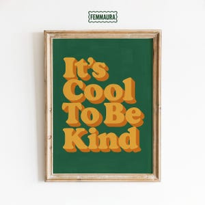 May include: Framed art print with the text "It's Cool To Be Kind" in orange, 3D-style lettering on a dark green background. The print is in a wooden frame and features the logo "FEMMAURA" at the top.