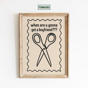 May include: Framed art print featuring the text "when are u gonna get a boyfriend???" above a black and white illustration of crossed scissors. The print has a beige background and a decorative wavy border, set in a light wood frame.