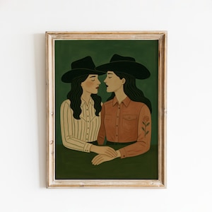 May include: A framed art print featuring two women in cowboy hats, facing each other. One wears a striped shirt, the other a rust-colored shirt with a rose tattoo. The background is a dark green, and the frame is a light wood color.
