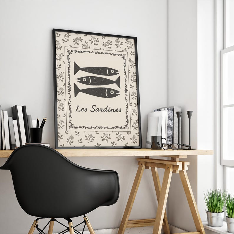May include: A framed art print featuring three stylized black fish above the words "Les Sardines." The print has a cream-colored background with a floral pattern and a black frame. The artwork is displayed on a wooden desk.