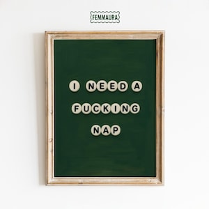 May include: A framed green board with the text "I NEED A FUCKING NAP" in white letter tiles. The frame is a light brown color. The logo "FEMMAURA" is at the top of the image.