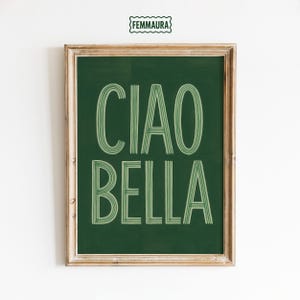 Op de afbeelding: Een ingelijste kunstdruk met de woorden "CIAO BELLA" in lichtgroene, gestileerde tekst op een donkergroene achtergrond. De lijst is lichtbruin van kleur. De linkerbovenhoek van de afbeelding heeft de tekst "FEMMAURA" in een groene, golvende letter.