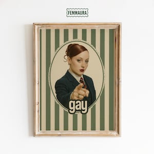 May include: Framed art print featuring a vintage-style illustration of a woman in a suit pointing at the viewer. The word "gay" is displayed in a bold font at the bottom. The background has green and white vertical stripes.