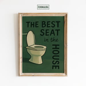 May include: Framed art print with a green background and a vintage-style illustration of a toilet. The text reads "THE BEST SEAT in the HOUSE" in black lettering. The frame is a light brown color.