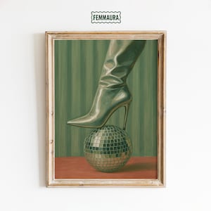 May include: Framed artwork featuring a high-heeled boot balancing on a disco ball. The boot is a metallic green color, and the disco ball is silver. The background has green vertical stripes, and the frame is a light brown color. The artwork is signed with the text "FEMMAURA".