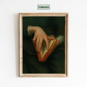 May include: Framed vintage art print featuring a person's hand holding an open book with a red cover. The artwork has a dark, moody aesthetic with a green and brown color palette. The print includes the text "FEMMAURA" at the top.