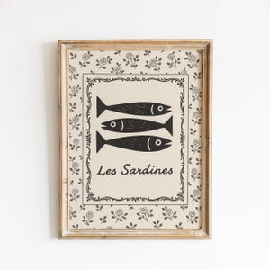 May include: Framed art print featuring three stylized black fish on a beige background, with the text "Les Sardines" below. The print is surrounded by a decorative border and a floral pattern, all within a wooden frame.