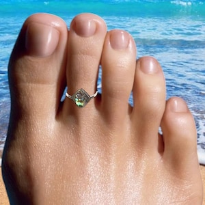 May include: A silver toe ring featuring a square, iridescent stone. The ring is worn on a foot, with a beach scene in the background. The stone displays green, blue, and purple hues.