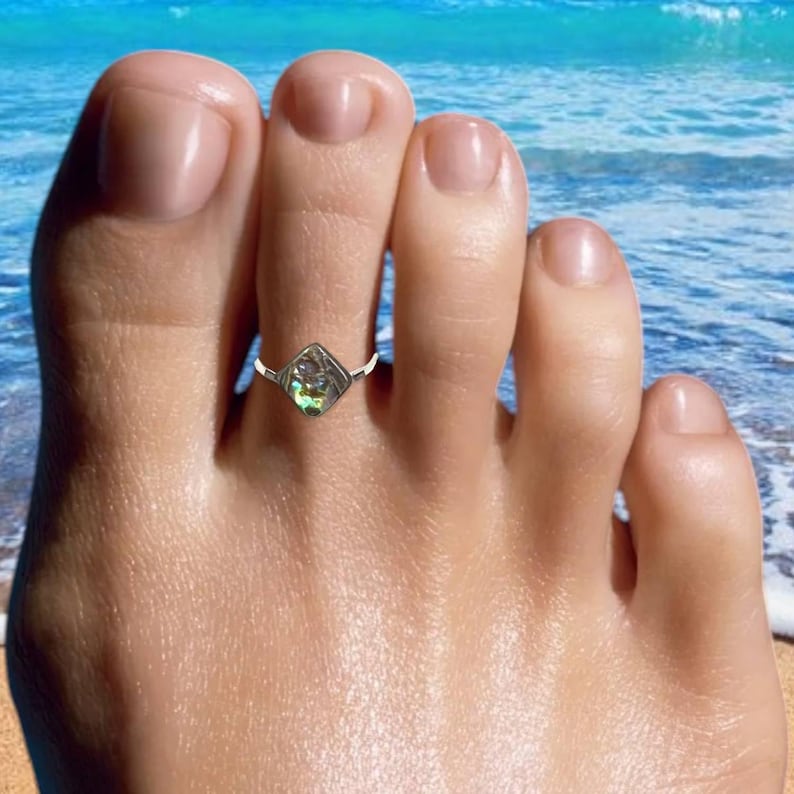 May include: A silver toe ring featuring a square, iridescent stone. The ring is worn on a foot, with a beach scene in the background. The stone displays green, blue, and purple hues.