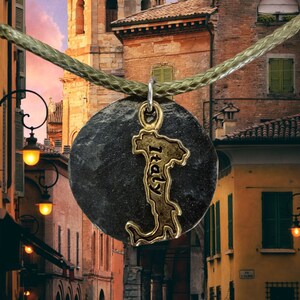 May include: A silver necklace with a round, dark gray pendant featuring a gold outline of the Italian peninsula. The pendant is hanging in front of a brick building with a terracotta roof. The building has a tower with a bell tower and a window with a green shutter. The background is a sunset sky.