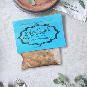 May include: A blue and white label with the text "Aunt Carol's Gourmet Dips" and "Chocolate Peanut Butter". The label is on a brown paper bag filled with a brown substance. The text "LOVE is the best ingredient" is written on a white cloth in the background.
