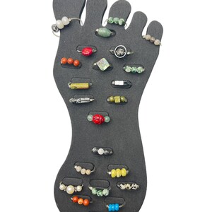 May include: A black foot-shaped display with 20 toe rings. The rings are made of silver and feature various colored beads, including red, green, white, black, and blue.