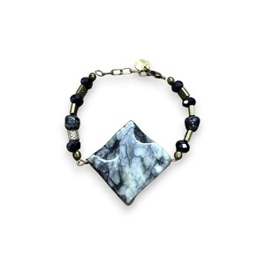 May include: A bracelet featuring a large, diamond-shaped, marbled stone in shades of gray and green. The bracelet includes black beads and gold-toned metal accents. The design is completed with a delicate chain and a small, round gold-toned clasp.