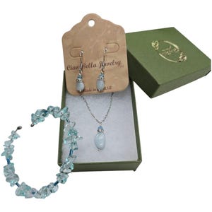 May include: A jewelry set featuring a necklace, earrings, and bracelet. The necklace and earrings have a light blue stone pendant and accents. The bracelet is made of light blue stones. The set is displayed in a green box with a matching gift box.