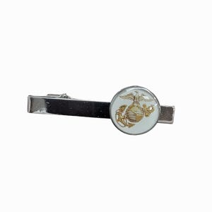 May include: A silver-toned tie clip with a circular emblem featuring a gold-colored eagle, globe, and anchor design on a white background. The clip is rectangular and has a clasp on the back.