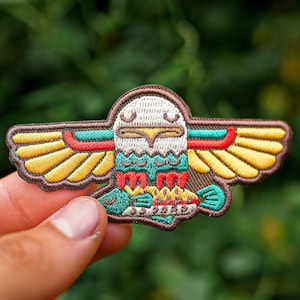 May include: Embroidered patch featuring a stylized eagle design with outstretched wings. The patch has a brown border, with yellow, red, and teal accents. The eagle's head is white, and the wings are yellow with red and teal details.