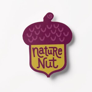 May include: A sticker in the shape of an acorn with a purple top and a yellow base. The text "nature nut" is written in a whimsical font on the yellow base. The sticker is on a white background.