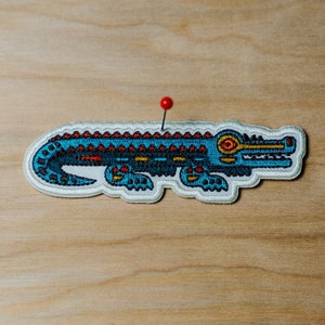 May include: Embroidered alligator patch in teal, red, and gold. The patch is outlined in white and has a red pin through it, set against a light wood grain background. The alligator design features intricate details.