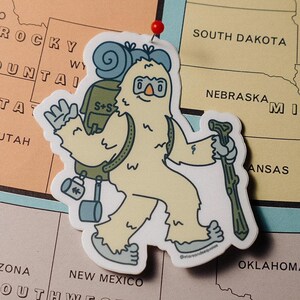 May include: A die-cut sticker of a cartoon yeti hiker with a backpack and walking stick. The yeti is light yellow with blue accents, and the sticker is placed on a map of the United States.