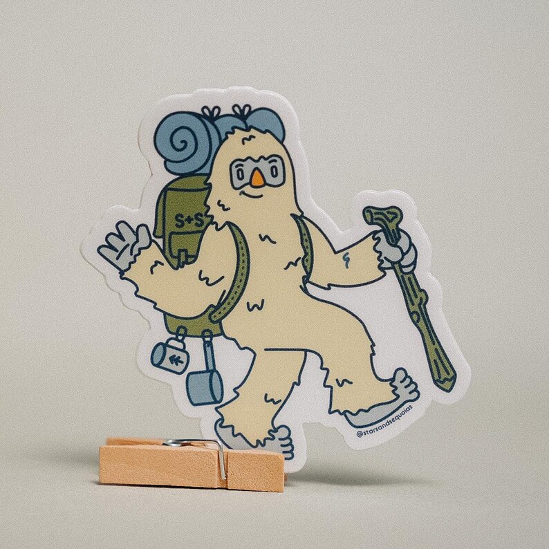 May include: A die-cut sticker featuring a cartoon Bigfoot character with a backpack and walking stick. The Bigfoot is light yellow with blue and green accents. The sticker has the text "S+S" on the backpack and "@starsandsequoias" at the bottom.
