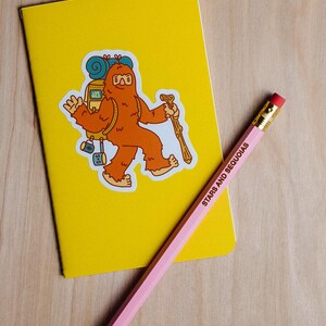 May include: A yellow notebook with a sticker of a cartoon Bigfoot carrying a backpack and a walking stick. A pink pencil with the text "STARS AND SEQUOIAS" lies diagonally across the notebook.