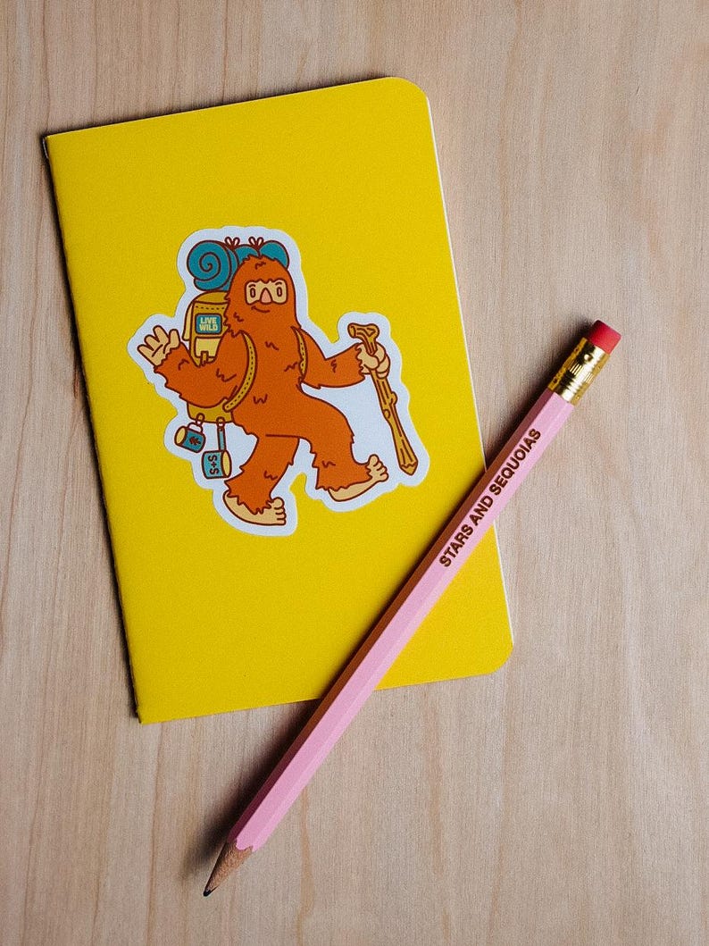 May include: A yellow notebook with a sticker of a cartoon Bigfoot carrying a backpack and a walking stick. A pink pencil with the text "STARS AND SEQUOIAS" lies diagonally across the notebook.