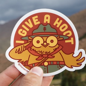 May include: A round sticker featuring a cartoon owl wearing a ranger hat and uniform. The owl has large yellow eyes and a red neckerchief. Text on the sticker reads "I GIVE A HOOT" in yellow letters. The sticker has a white border and the text "@starsandsr" at the bottom.