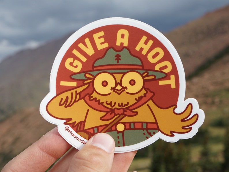 May include: A round sticker featuring a cartoon owl wearing a ranger hat and uniform. The owl has large yellow eyes and a red neckerchief. Text on the sticker reads "I GIVE A HOOT" in yellow letters. The sticker has a white border and the text "@starsandsr" at the bottom.