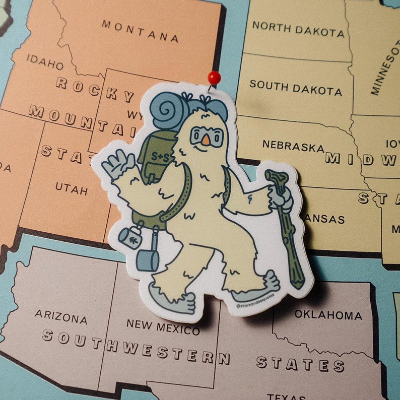 May include: A die-cut sticker of a cartoon yeti hiker with a backpack and walking stick. The yeti is light yellow with blue accents, and the sticker is placed on a map of the United States.