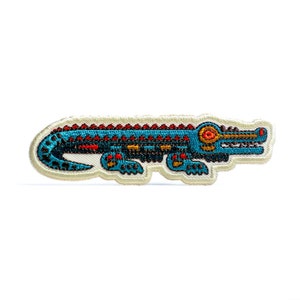 May include: Embroidered patch featuring a stylized alligator design. The alligator is primarily teal with red and black accents, outlined in black, and set against a cream-colored background. The patch is rectangular with a scalloped edge.