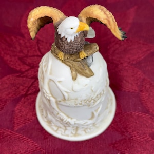 Puede incluir: Campana de cerámica con un águila calva detallada posada en una rama. El águila tiene plumas marrones y blancas, un pico amarillo y alas extendidas. La campana es blanca con una superficie texturizada y un diseño decorativo.