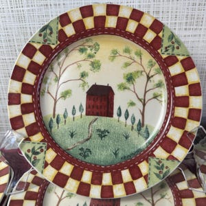 May include: A decorative plate with a rural landscape scene. The plate features a red brick house on a green hill, surrounded by trees. The rim has a red and yellow checkered pattern with green and red floral accents.